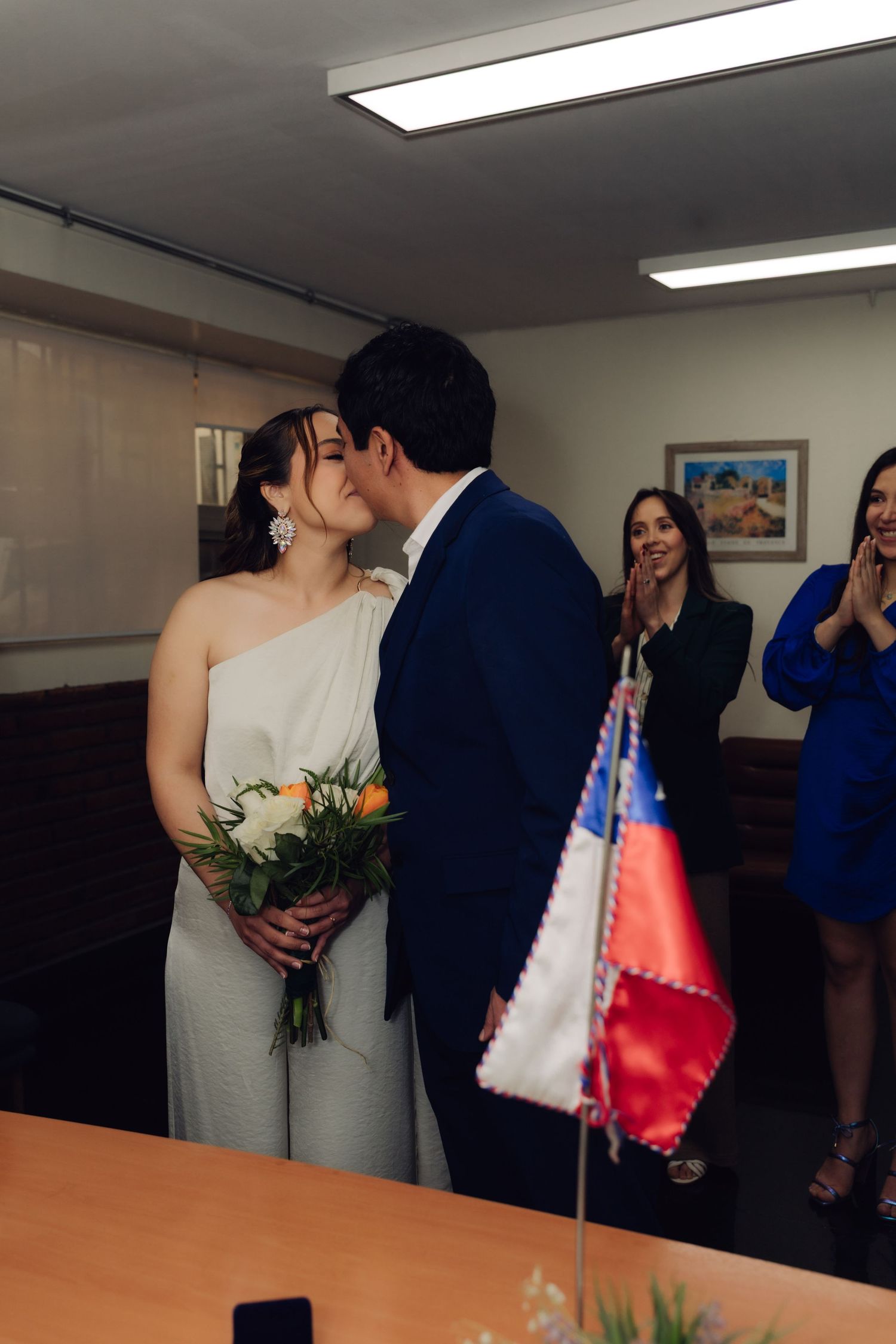 Newlyweds share first kiss at civil ceremony as guests applaud beside Chilean flag.