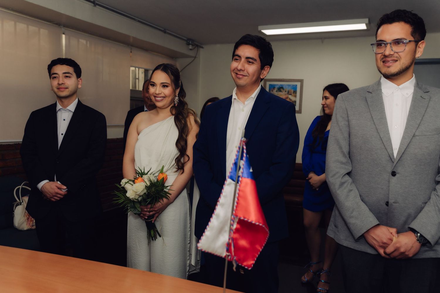 Wedding party smiles beside Chilean flag during civil marriage ceremony indoors.