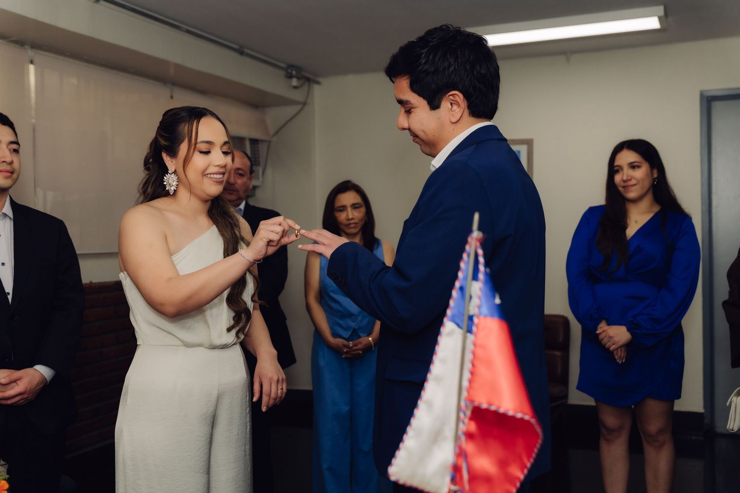 Couple exchanging rings during a civil wedding ceremony with Chilean flag and guests in background.