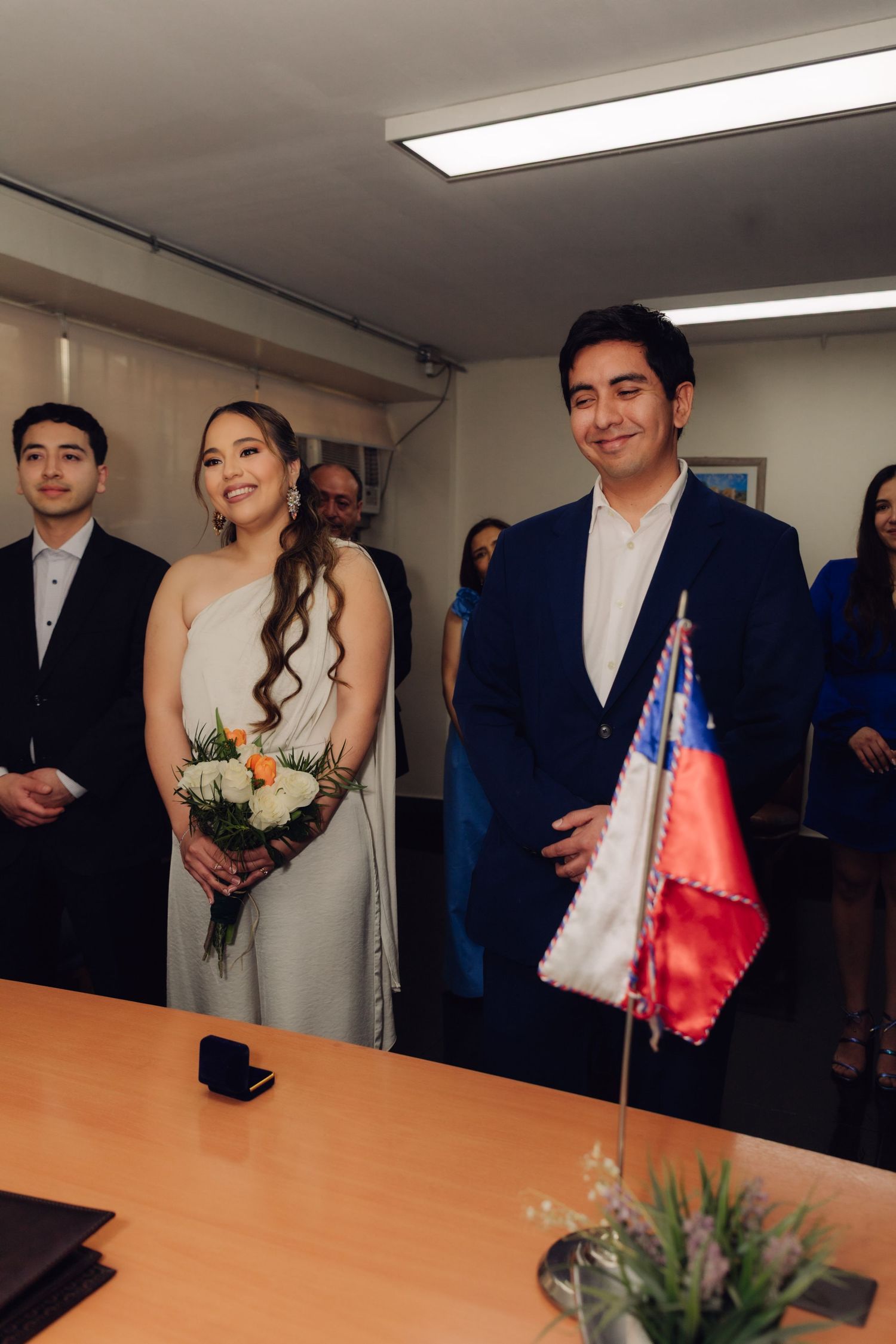 Bride and groom standing at civil ceremony table with Chilean flag, bride holding bouquet.