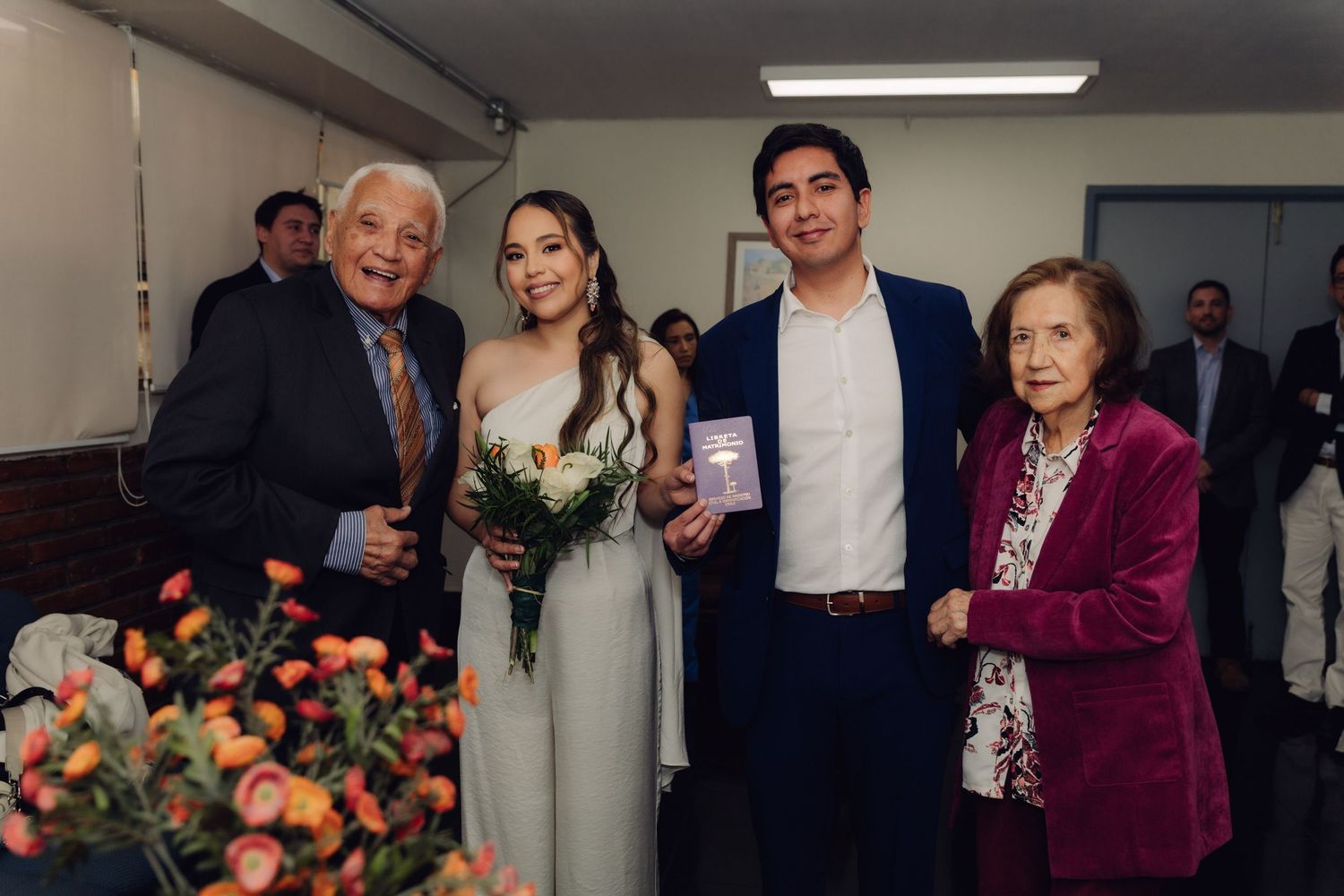 Newlyweds posing with elderly grandparents holding bouquet and marriage certificate after ceremony.