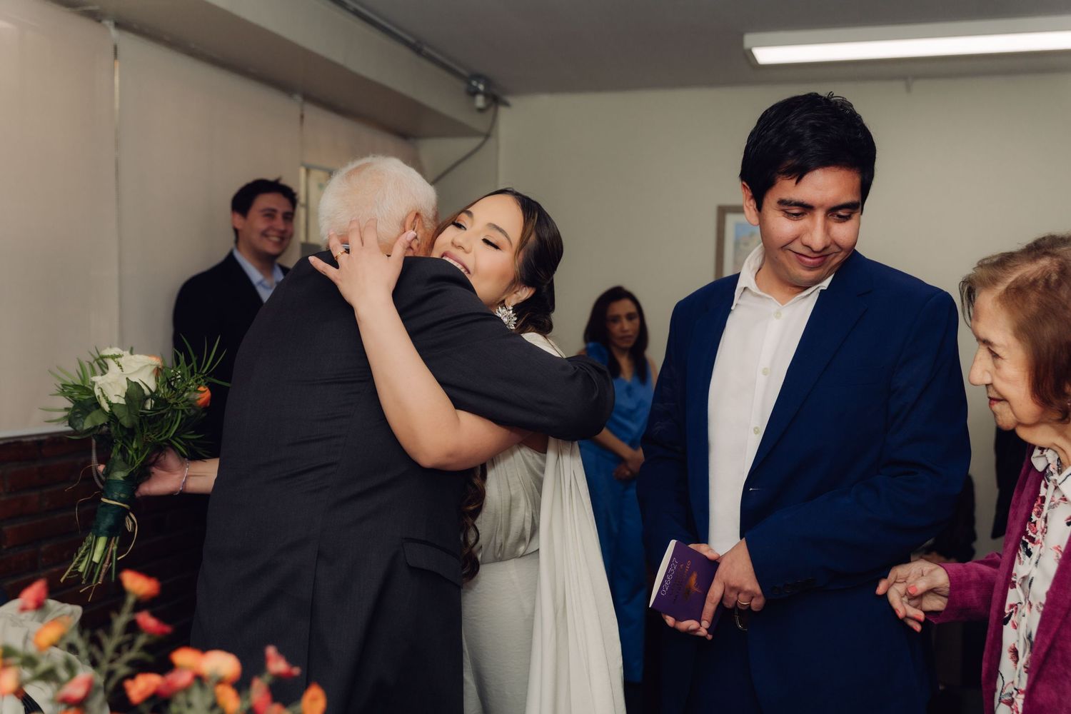 Bride embracing elderly guest while groom stands nearby after civil wedding ceremony.