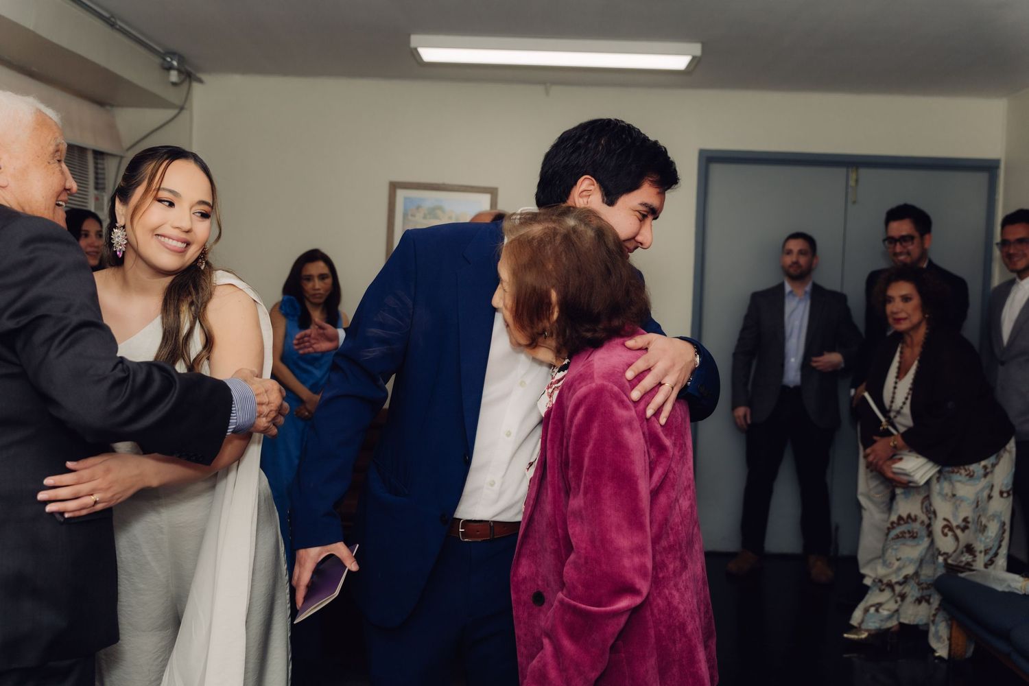 Groom hugging elderly woman in pink jacket while bride greets guests after wedding ceremony.