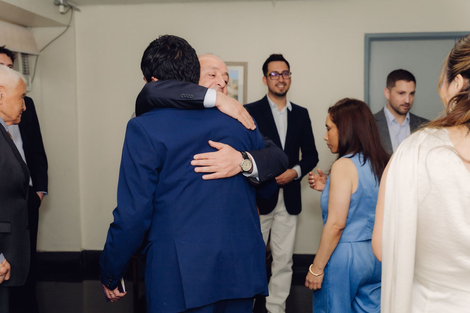 Wedding guests share warm hugs and congratulations in a bright indoor reception space.