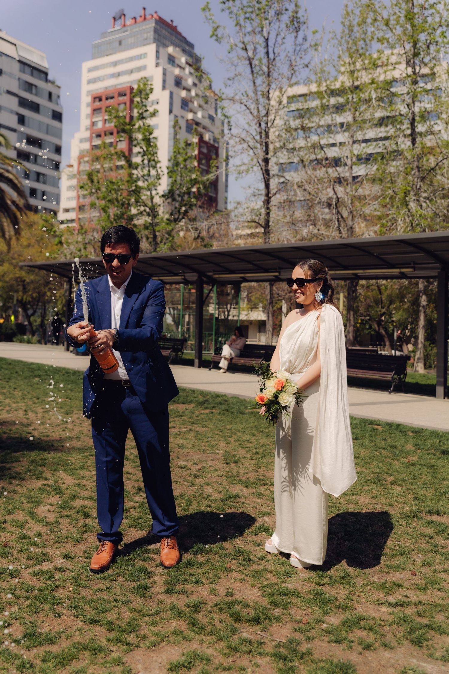 Bride and groom celebrating in urban park, groom popping champagne while bride holds flowers.