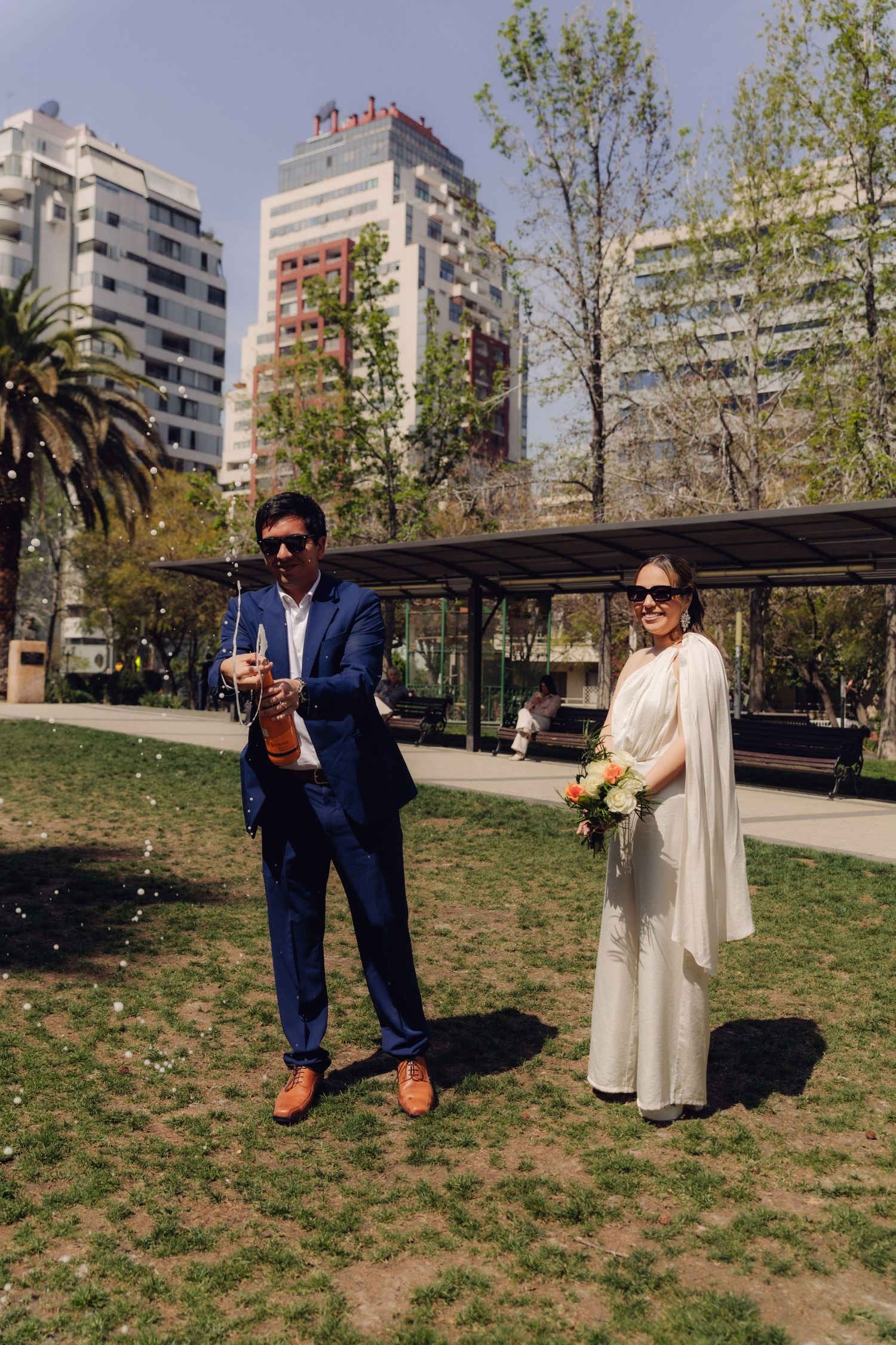 Newlyweds in a sunny park, groom in blue suit opening champagne bottle, bride holding bouquet.