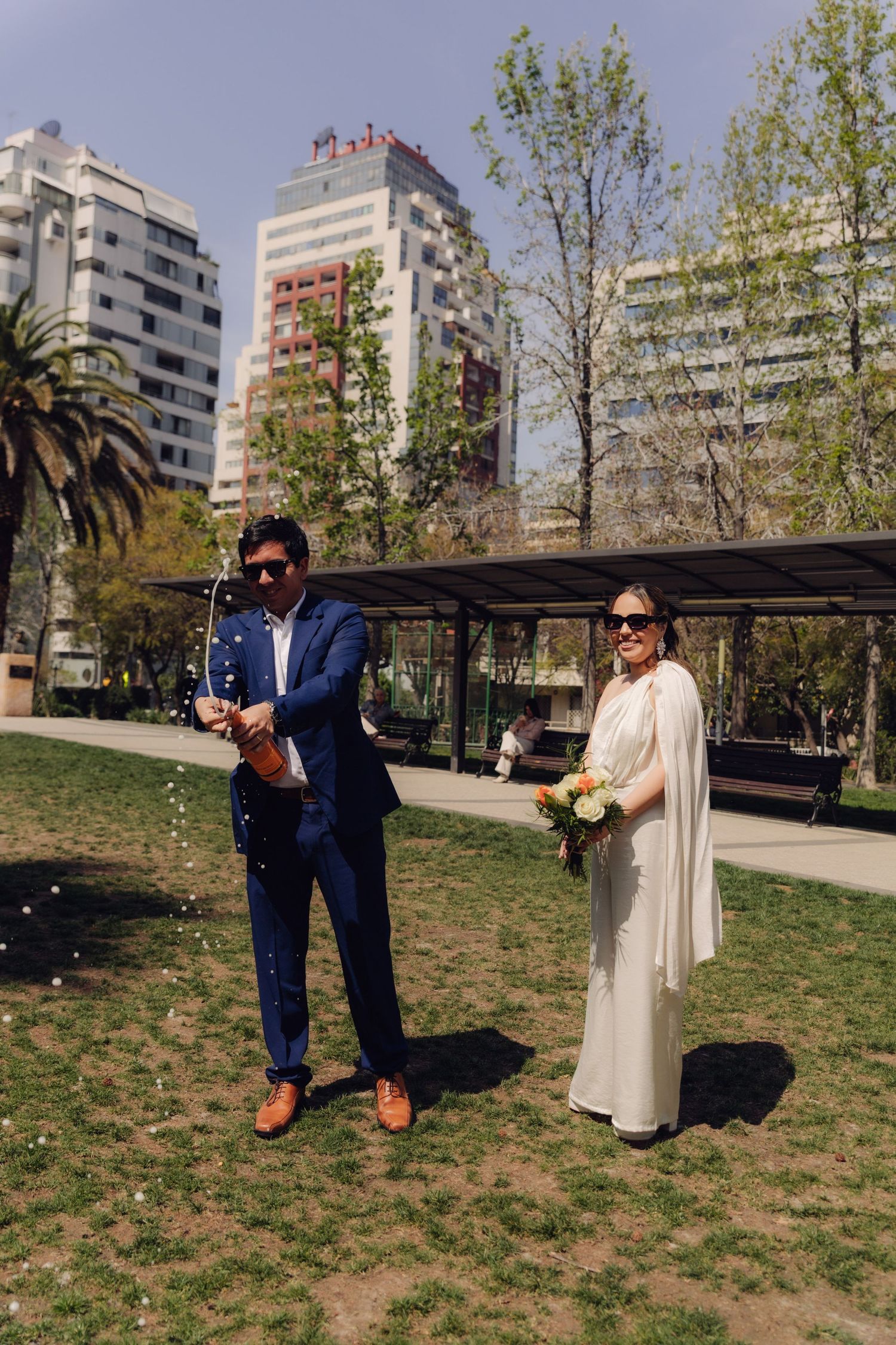 Wedding couple in sunglasses celebrating outdoors with champagne and bridal bouquet in city park.