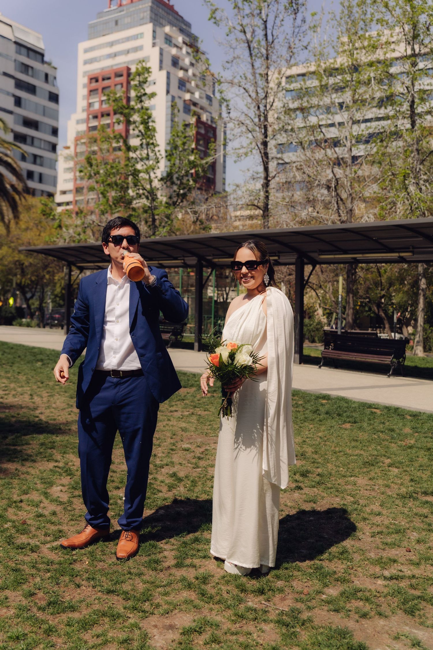 Groom in blue suit drinking champagne beside bride in white dress holding bouquet in sunny park.