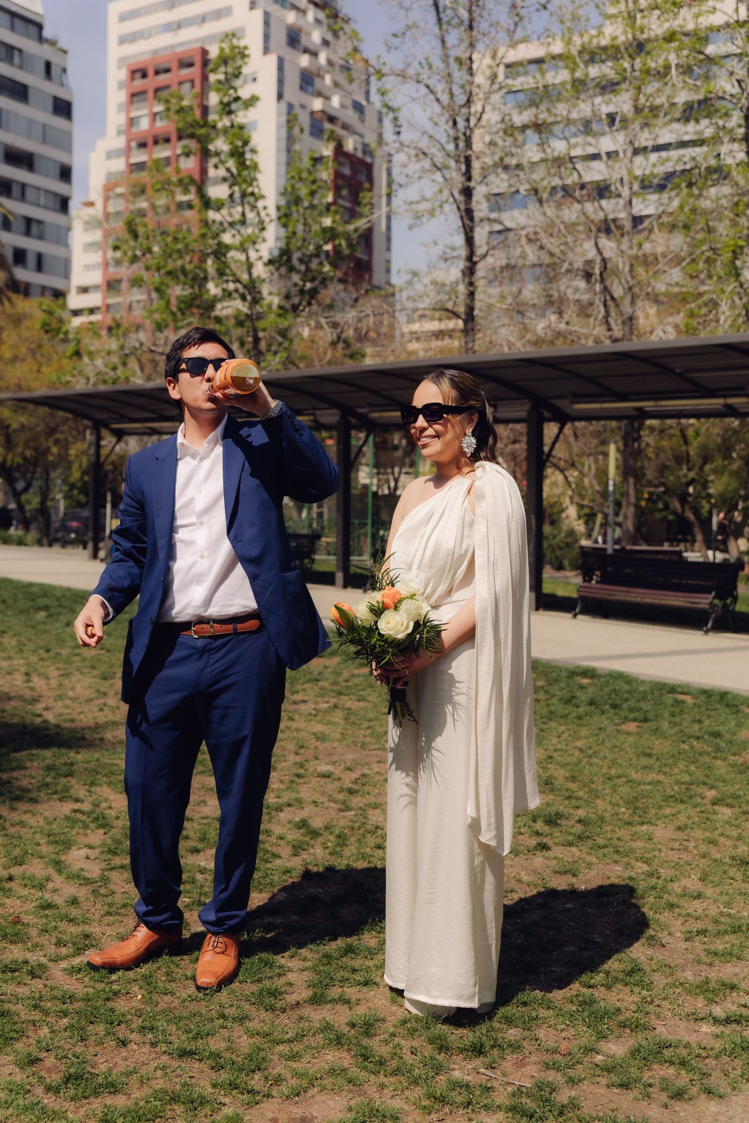 Smiling newlyweds in sunglasses, groom drinking champagne, bride holding bouquet in urban park.