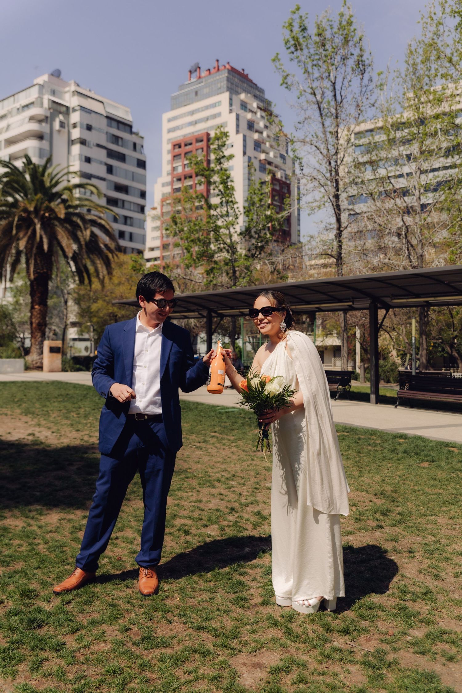 Bride and groom in sunglasses posing with champagne bottle in a sunny city park after wedding.