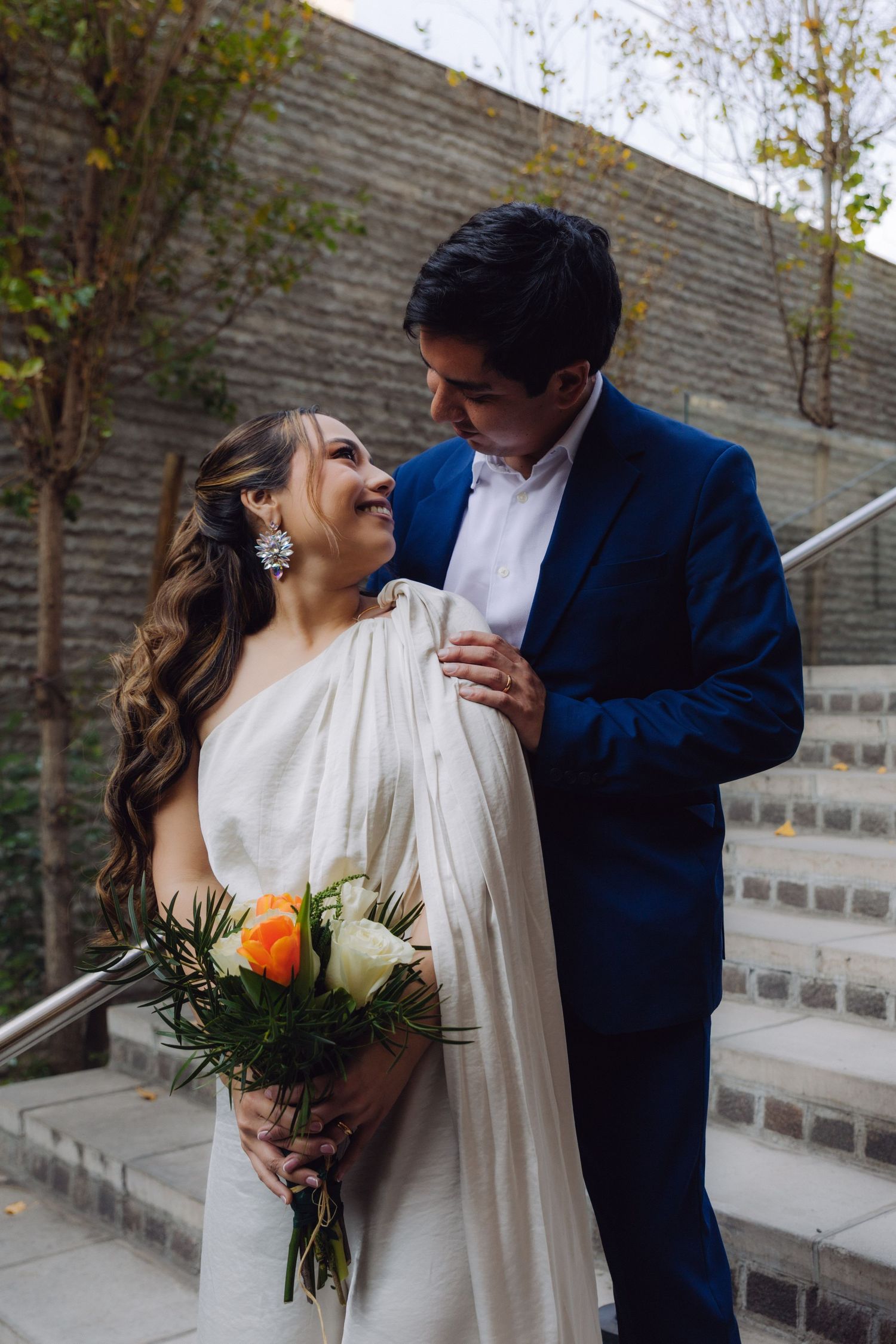 Romantic newlyweds in blue suit and white gown gazing at each other on outdoor stone steps.