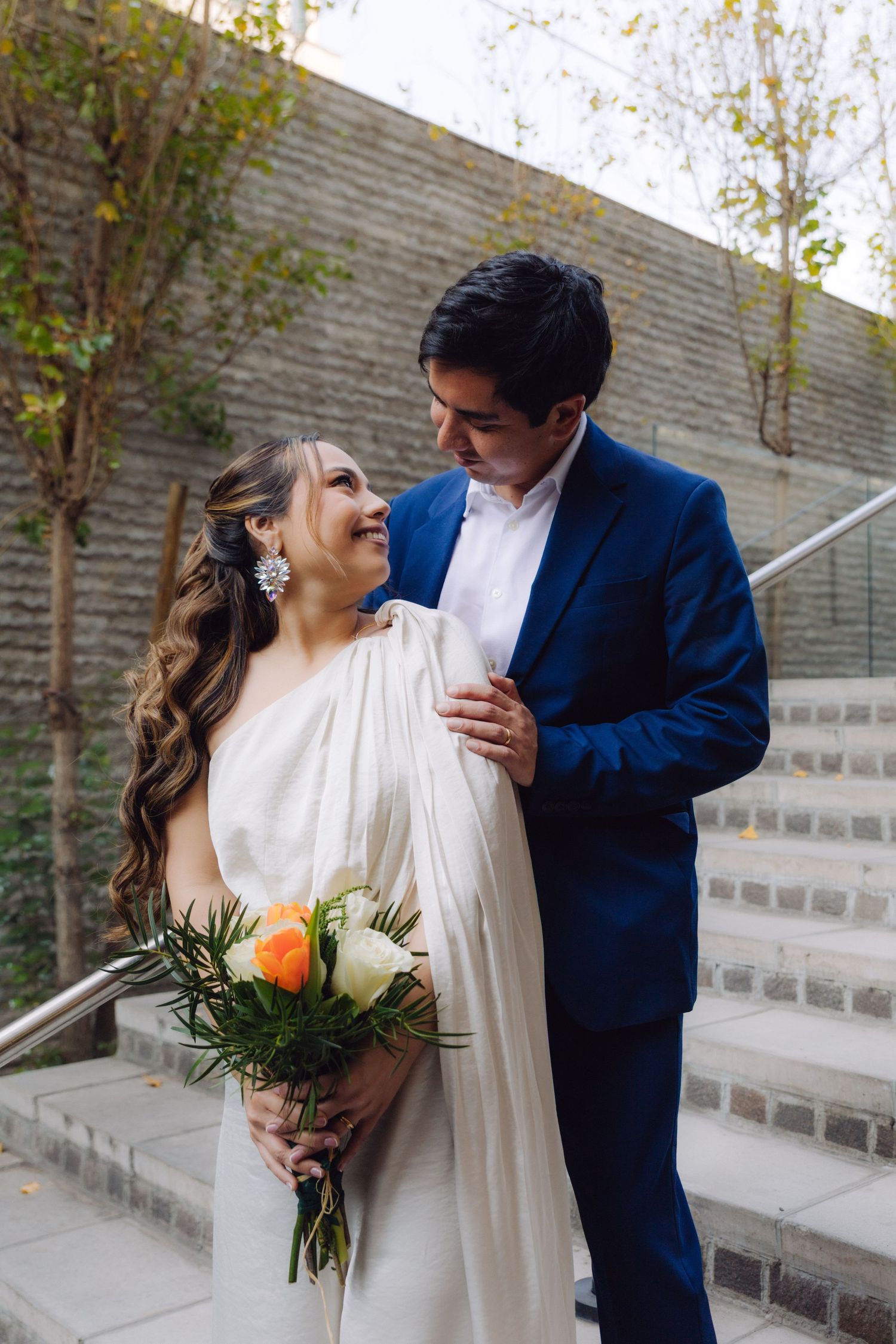 Bride in white dress holding colorful bouquet gazes at groom in blue suit on outdoor staircase.