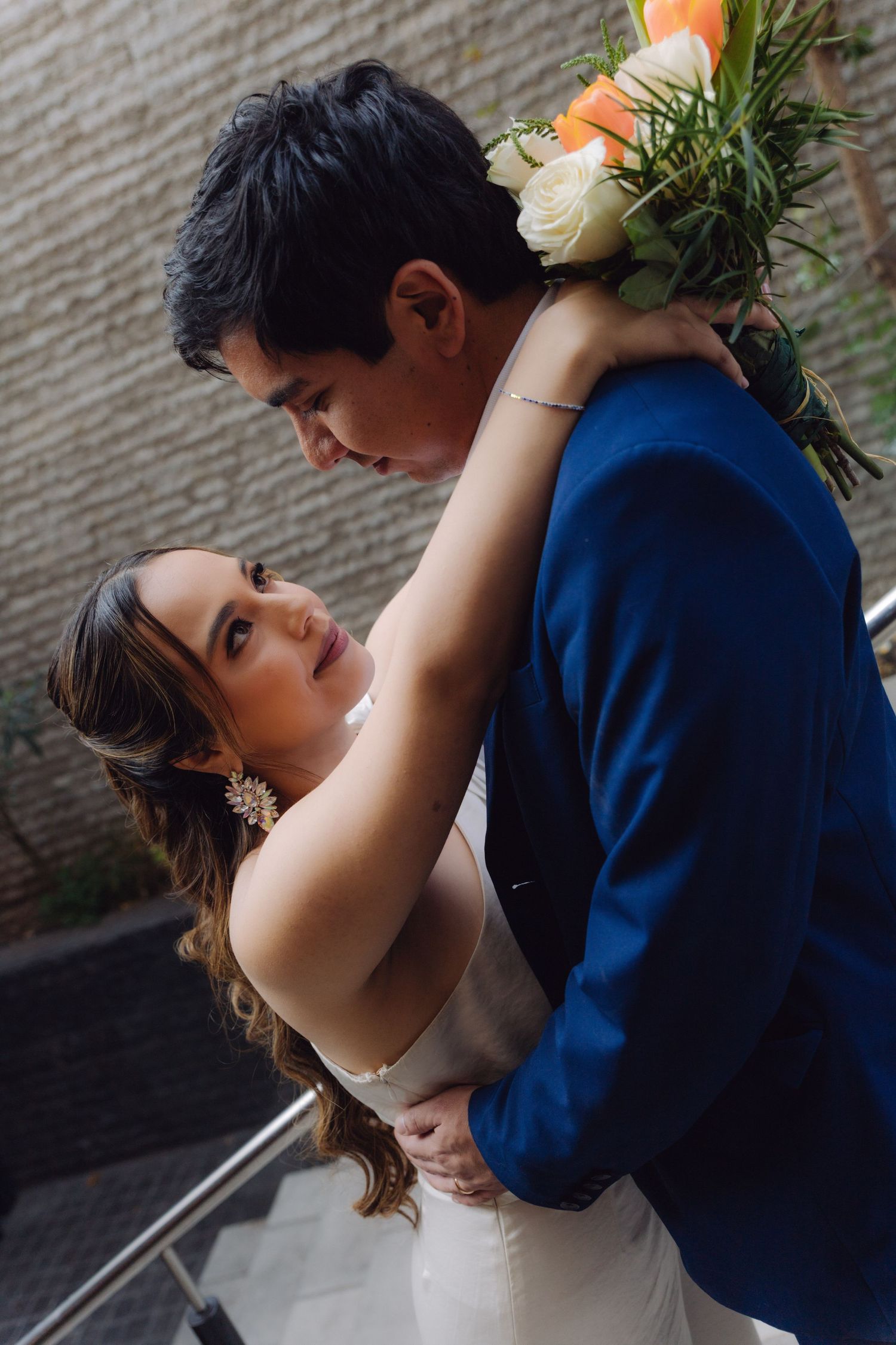 Bride embraces groom from behind holding bouquet while looking at camera on outdoor stairs.