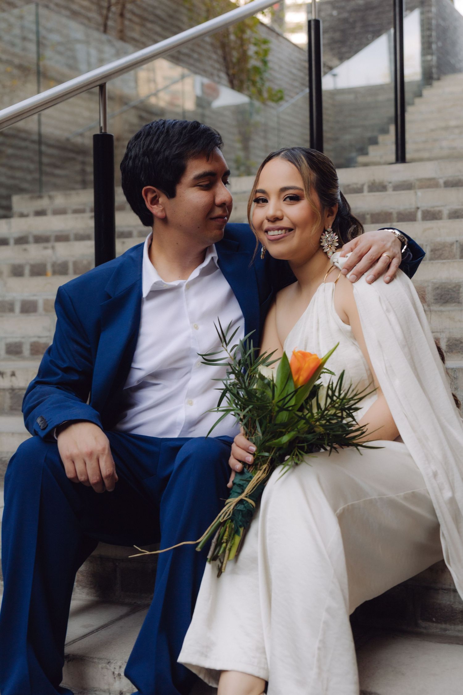 Smiling bride in white dress sits on steps with groom in blue suit arm around her shoulders.