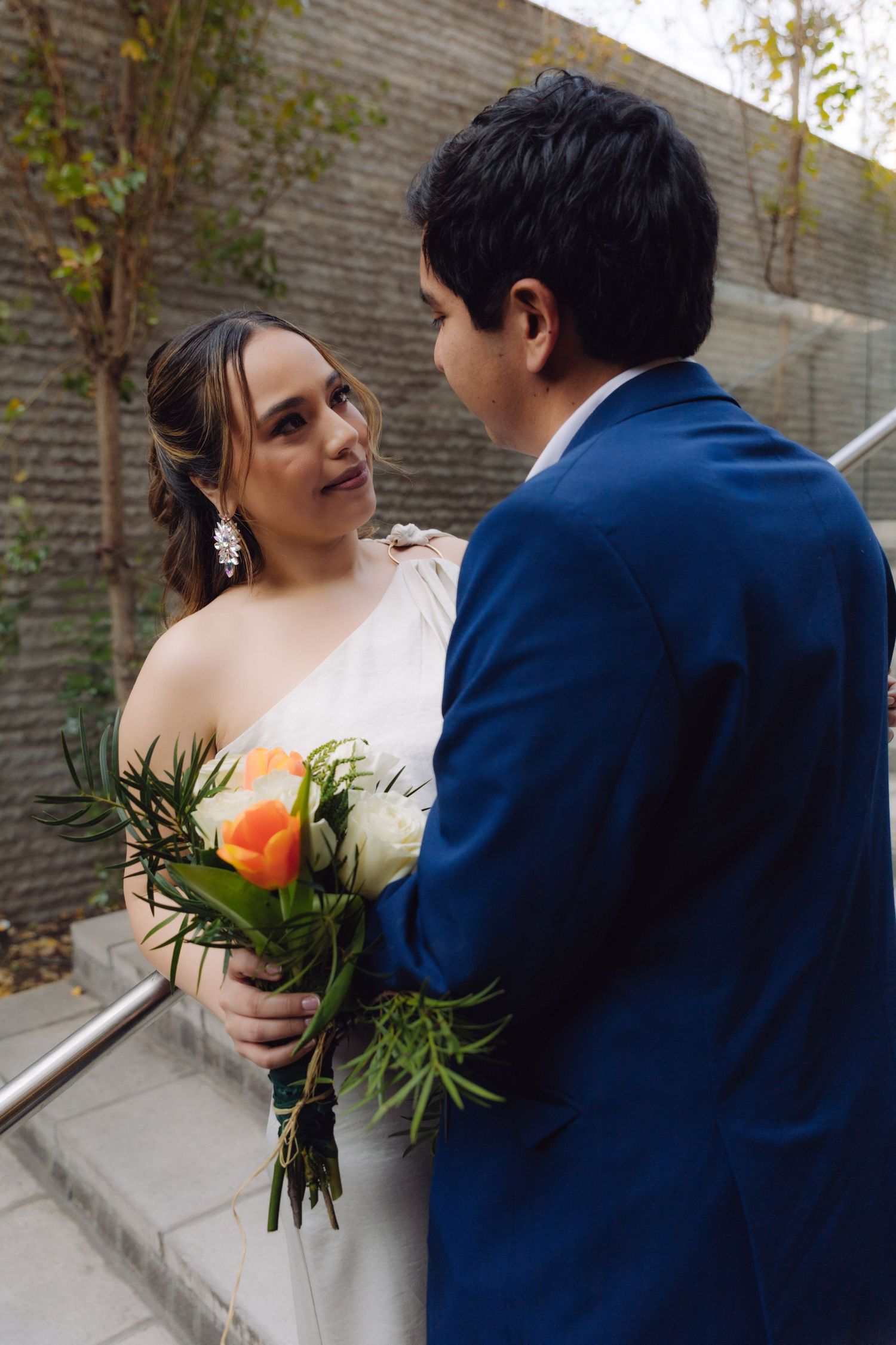 Bride holding orange and white bouquet looks up at groom in blue suit on outdoor staircase.