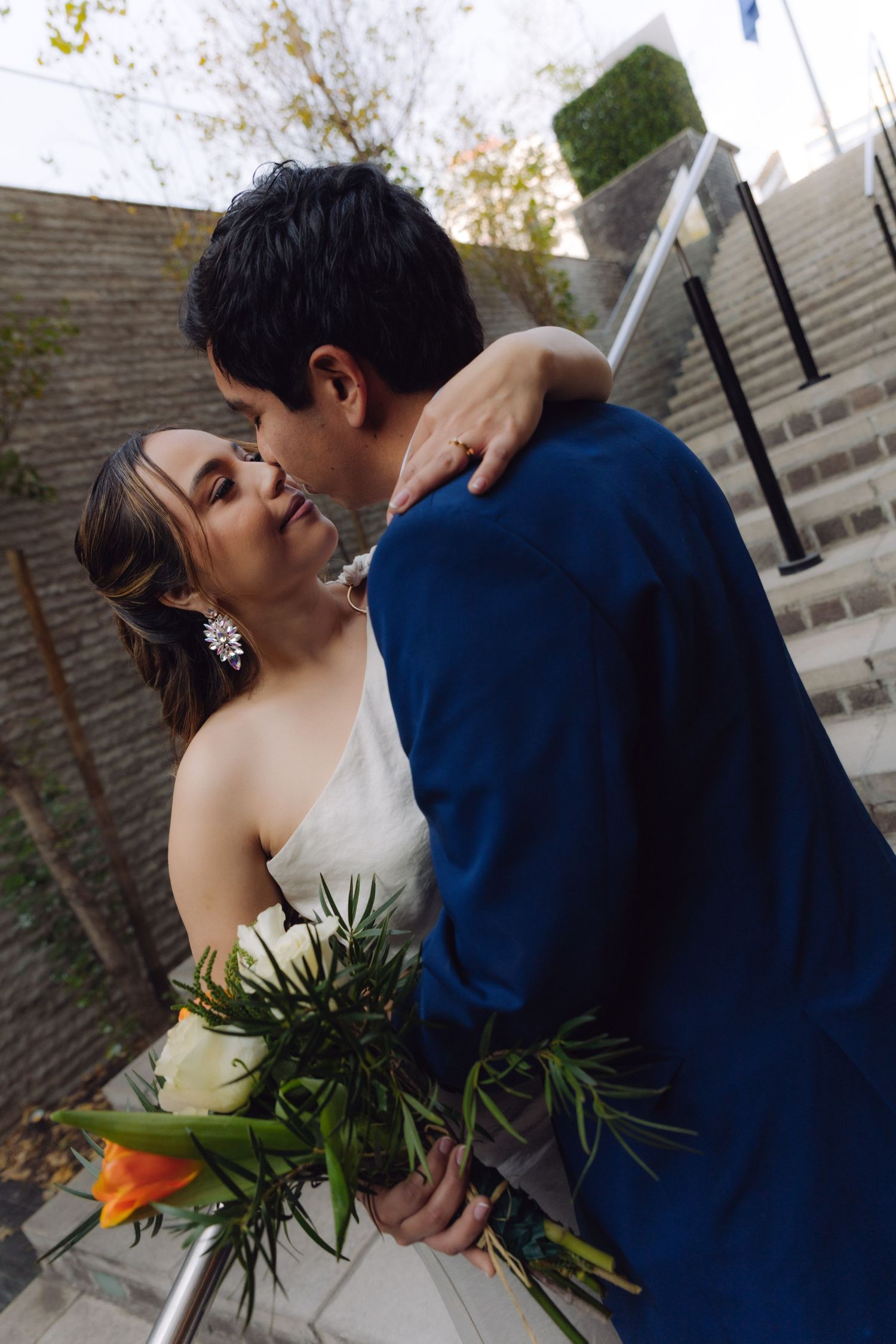 Bride and groom share a kiss on outdoor stone staircase, bride holding colorful floral bouquet.