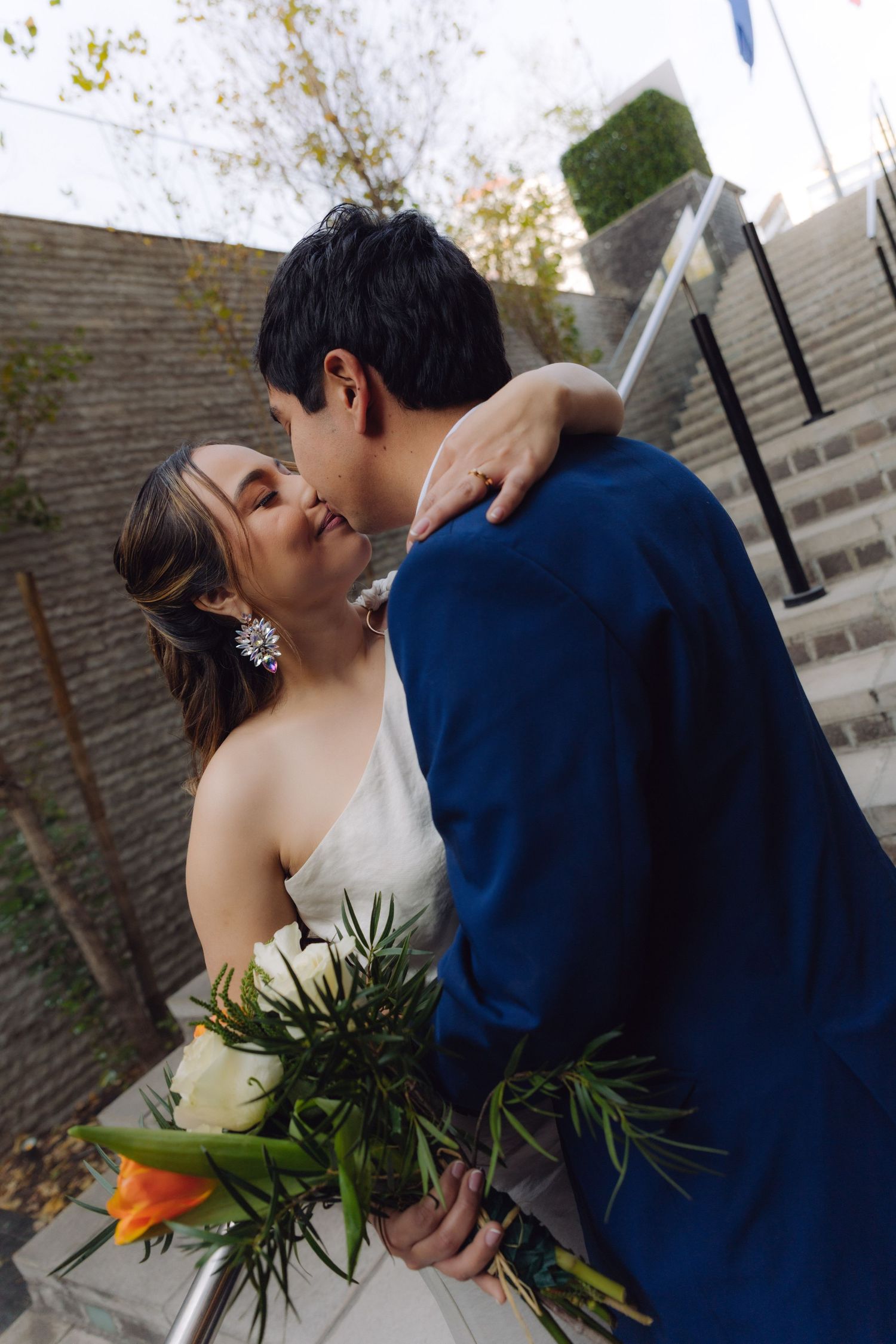 Wedding couple kissing on outdoor stairs, bride in white dress holding orange and white bouquet.