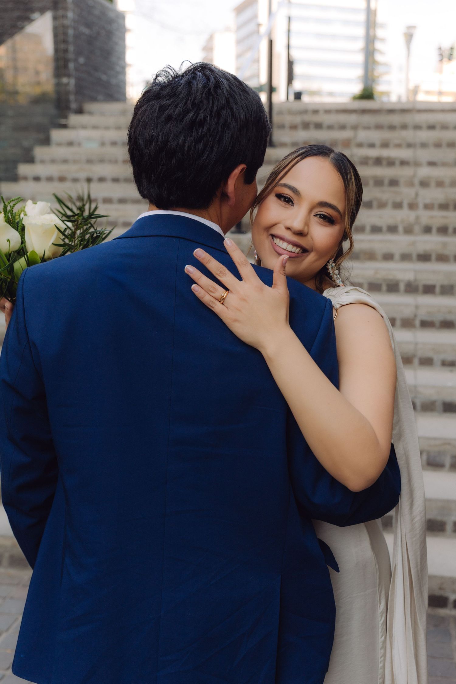 Smiling bride in white dress touches grooms face while embracing on outdoor staircase steps.