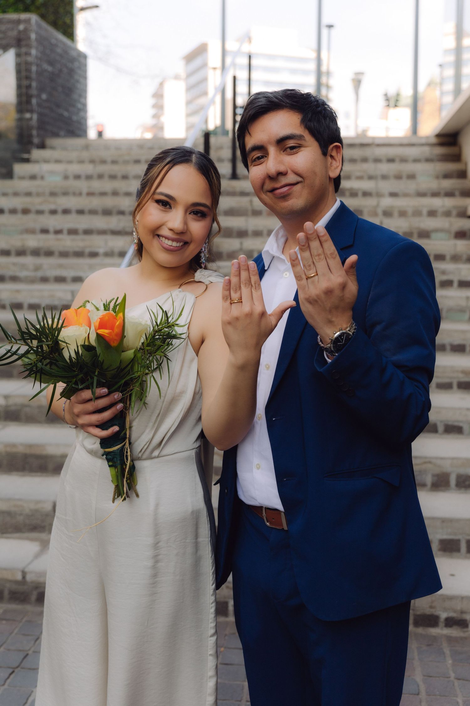 Newlywed couple smiling and showing wedding rings on outdoor staircase, bride holding bouquet.