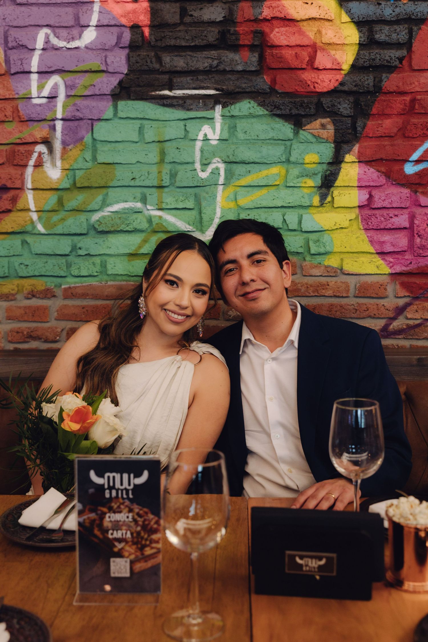 Newlywed couple smiling at camera seated at restaurant table with colorful graffiti wall behind.