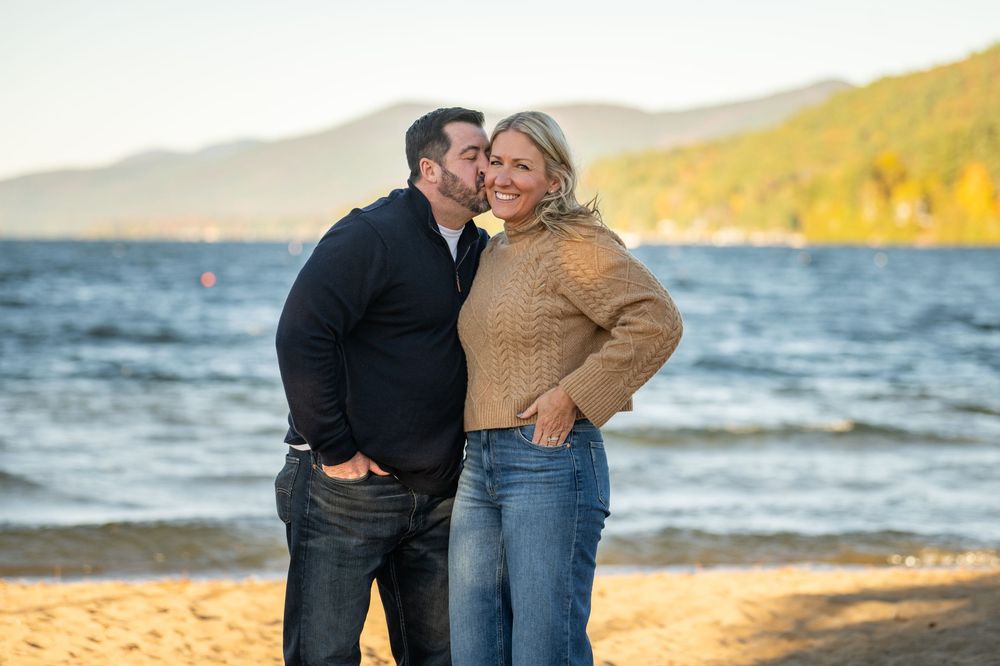 A couple shares an intimate moment on a beach at sunset with mountains and autumn foliage in the background.