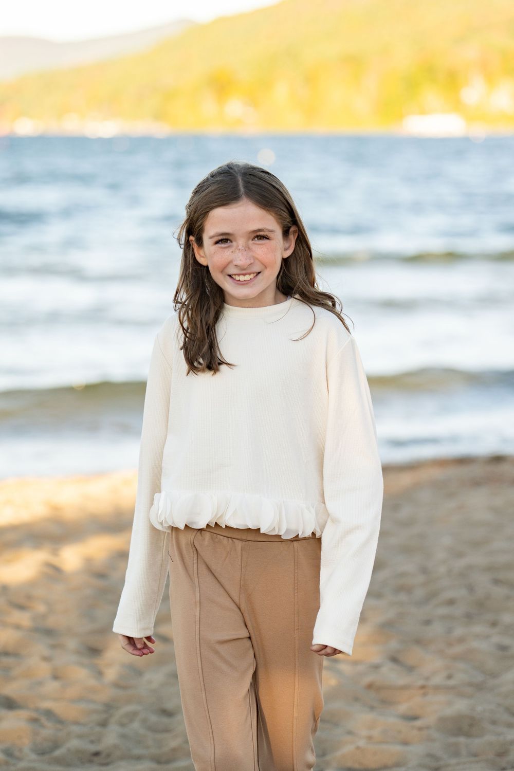 Person in a cream sweater and tan pants standing on a sandy beach during golden hour.