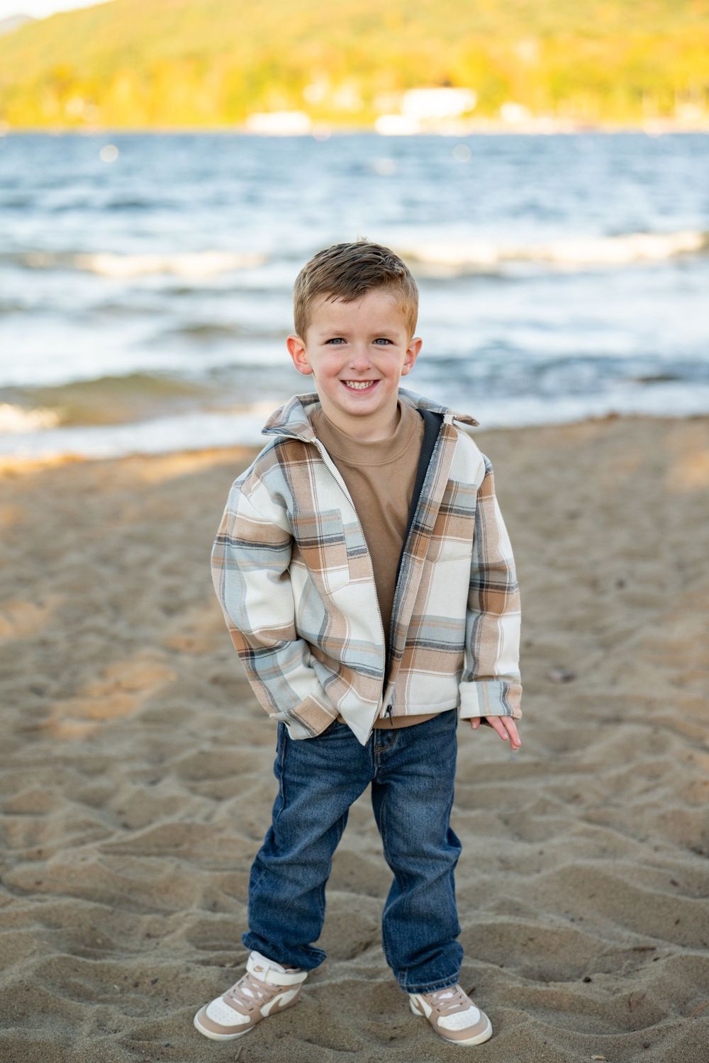 A young child in a plaid jacket and jeans plays on a sandy beach at sunset with ocean waves in the background.
