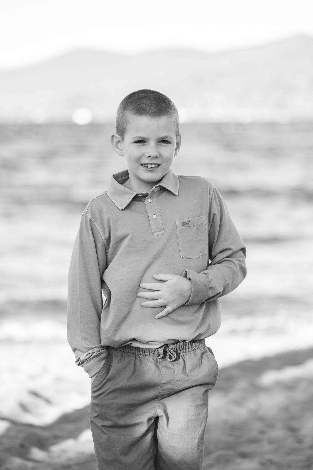 Black and white portrait of a young child in casual clothing standing outdoors by the water on a sunny day.