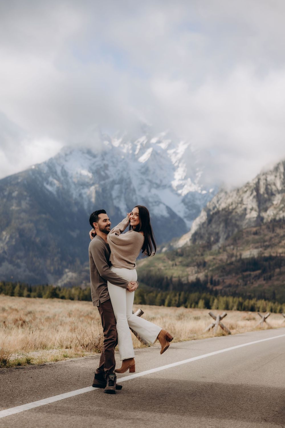 A couple dances playfully on a mountain road with snow-capped peaks and cloudy skies in the background.