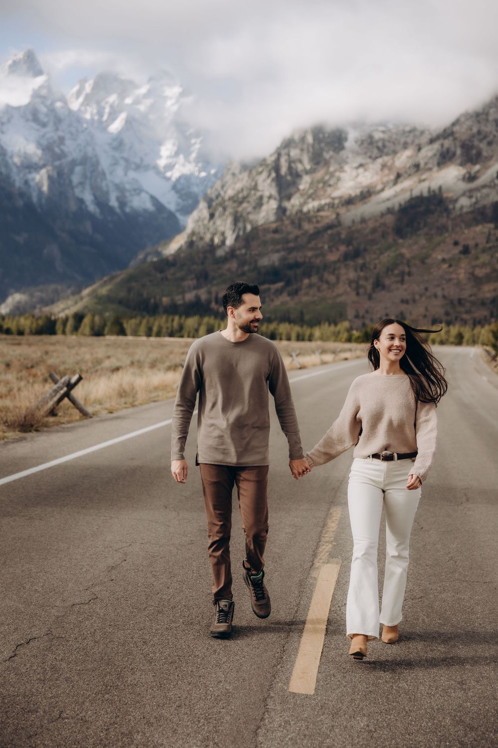 A couple walks playfully on a mountain road with snow-capped peaks and rugged terrain in the background.