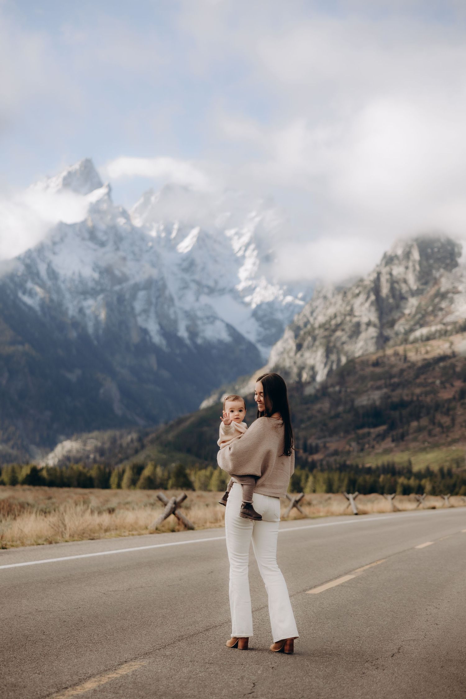 A couple embraces on an empty mountain road with snow-capped Grand Teton peaks rising dramatically in the background.