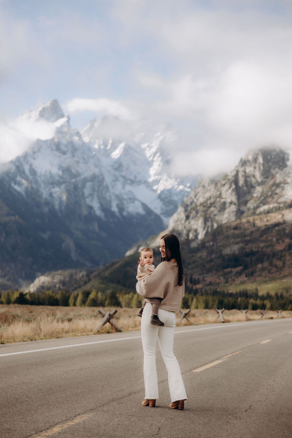 A couple embraces on an empty mountain road with snow-capped Grand Teton peaks rising dramatically in the background.