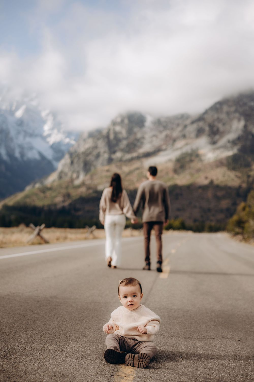 Baby sitting on empty mountain road while parents walk in distance with majestic mountain landscape behind.