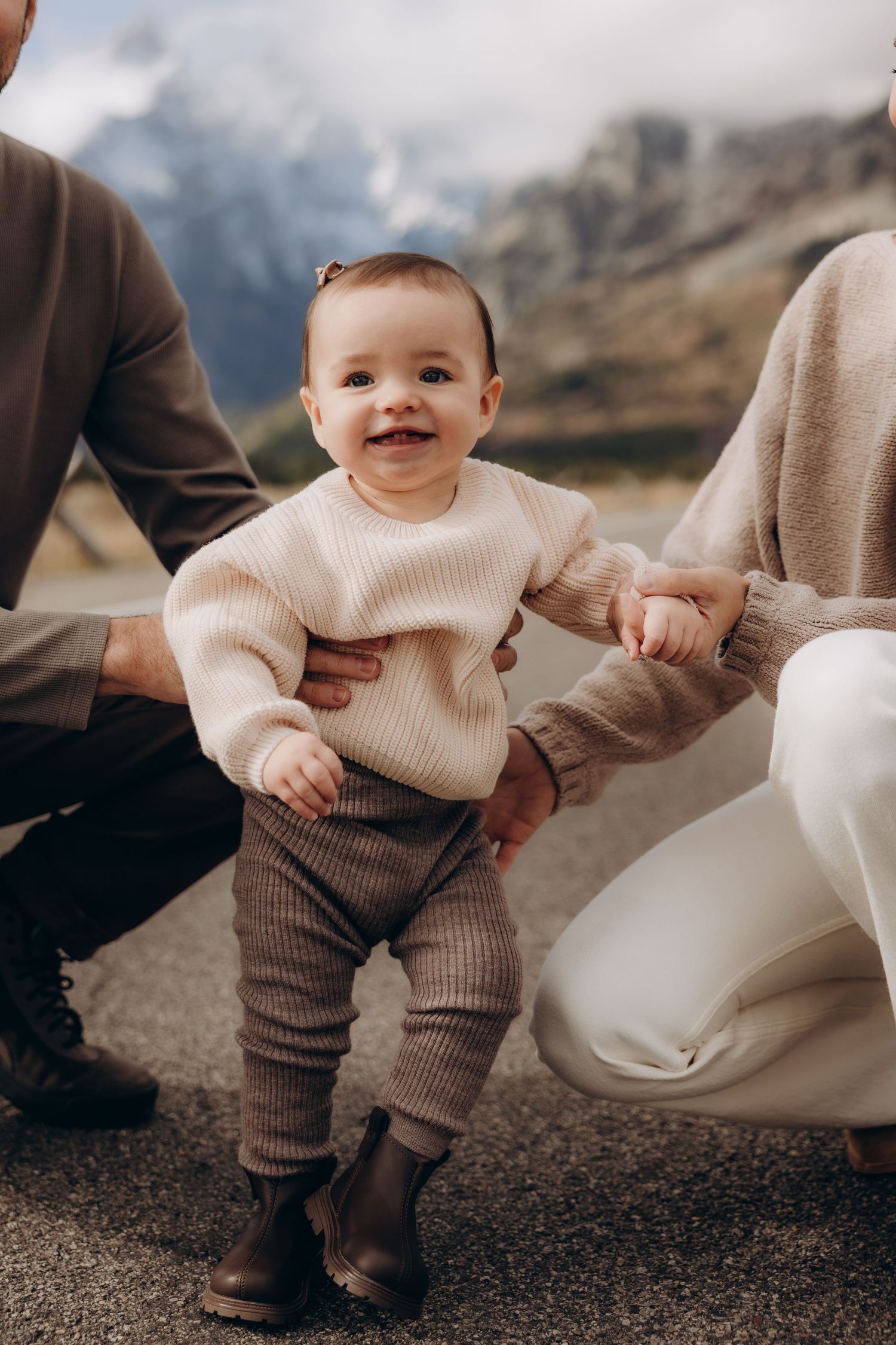A small child in a cream sweater and brown pants stands outdoors against a mountainous backdrop.