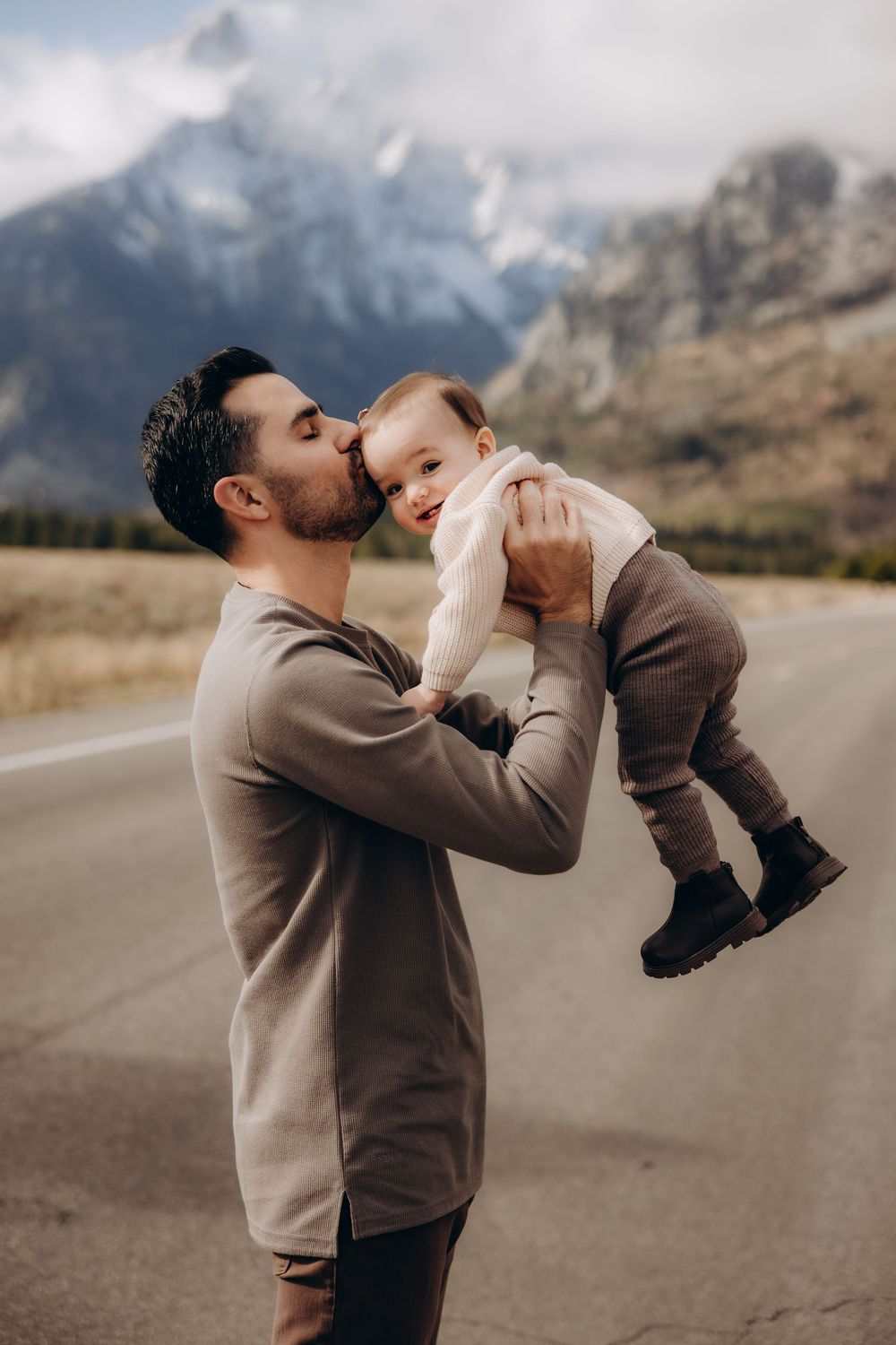 A parent in a beige sweater lifts and kisses a baby in a brown outfit against a mountainous backdrop on a road.