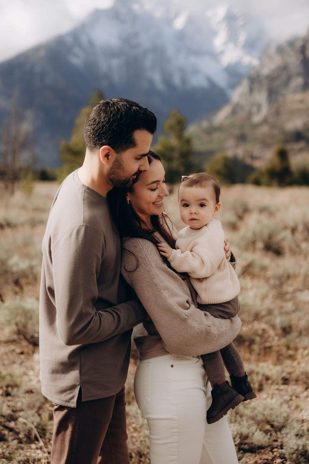 A family shares tender moments together against a backdrop of mountain scenery in neutral-toned clothing.