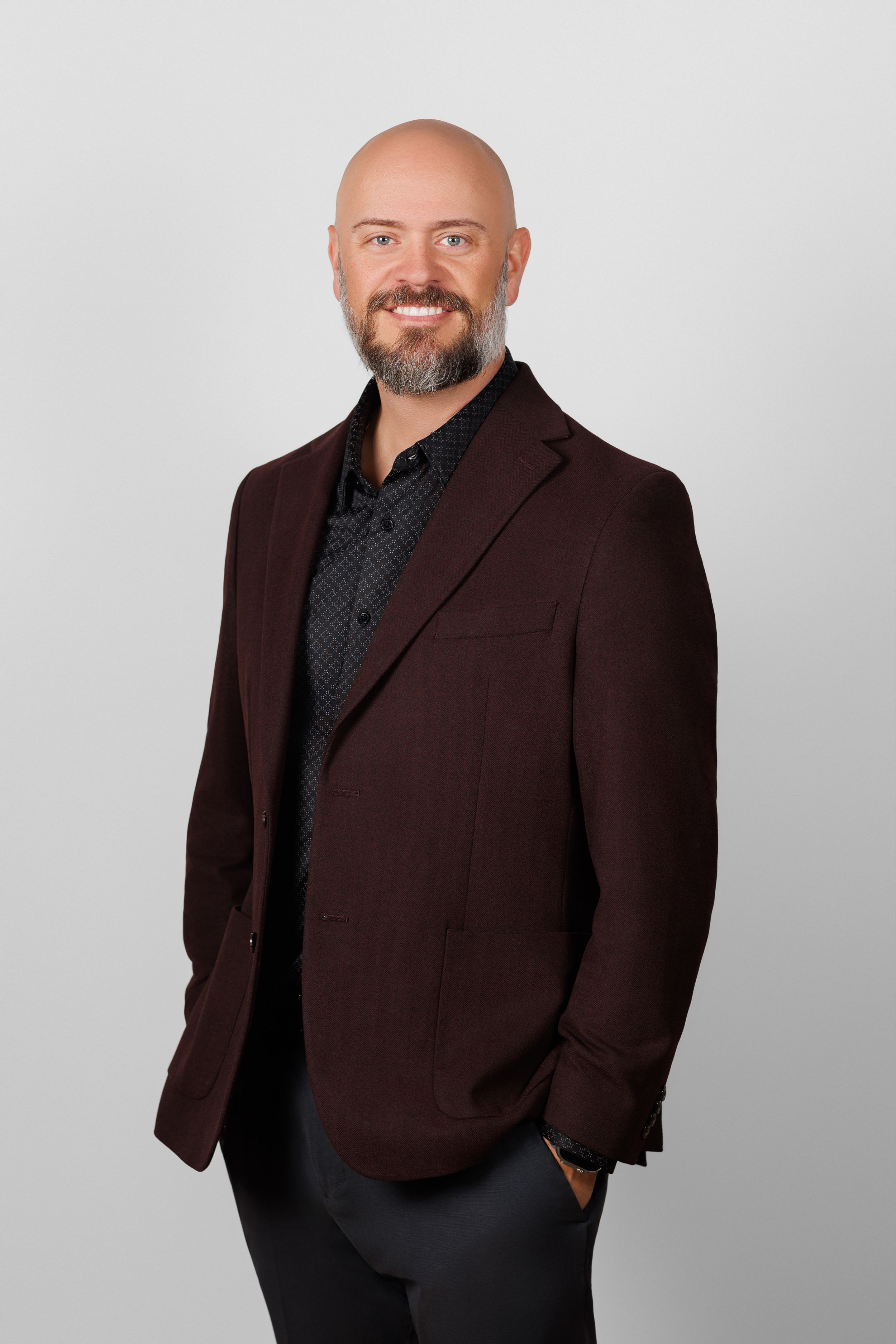 Professional headshot of a person in a brown blazer and dark shirt against a light gray background.