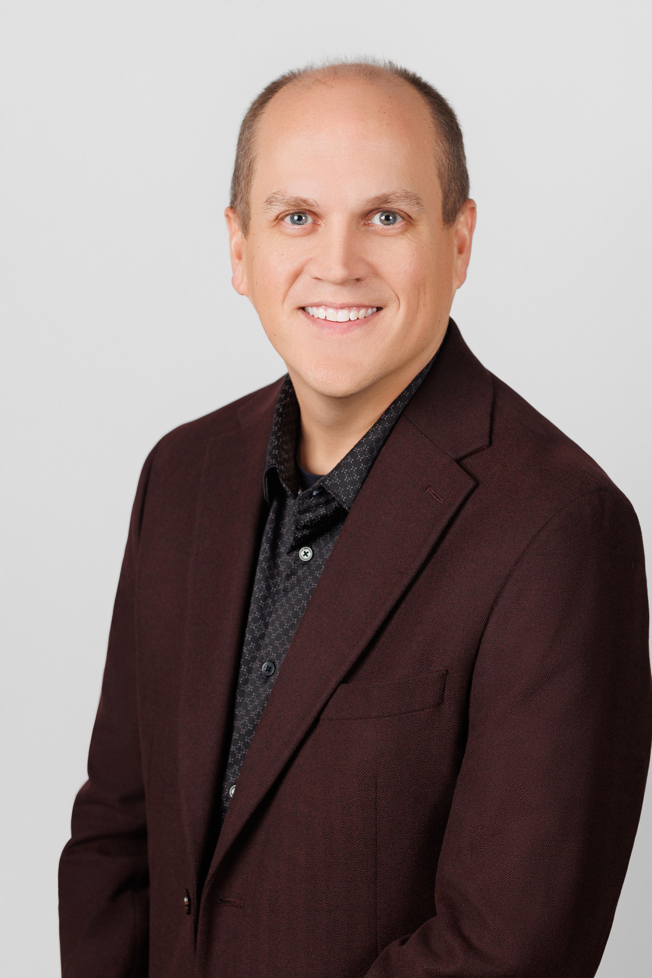 Professional headshot of a person wearing a brown suit jacket and black patterned shirt against a light background.