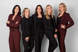 A group of five women pose together in coordinated black and burgundy business attire for a professional photo.