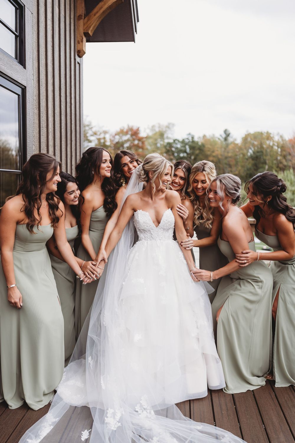 Bridesmaids in sage green dresses share a candid laughing moment with the bride in white on a wooden deck.