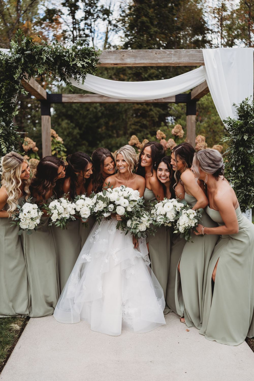 A bridal party in sage green dresses and white bouquets poses together for a candid laughing photo at an outdoor wedding.