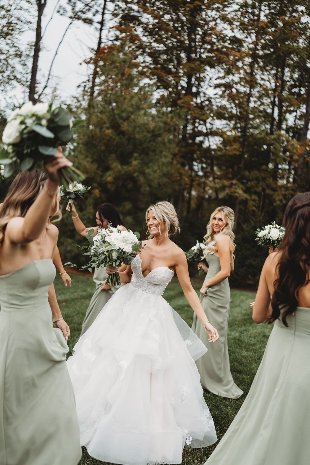 A bride in a white gown and bridesmaids in sage green dresses dance and celebrate joyfully outdoors.