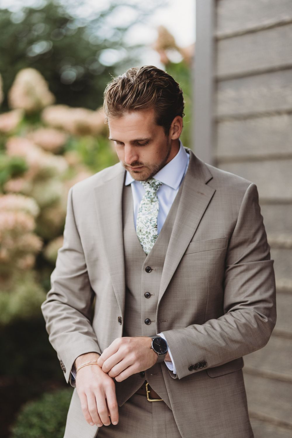 A groom in a light gray three-piece suit and floral tie adjusts his cuff links during wedding preparations.