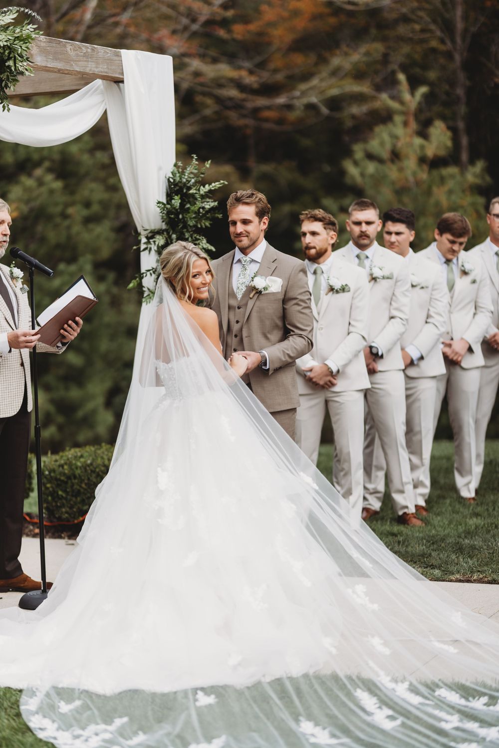 Elegant outdoor wedding ceremony with bridal party in white and grey attire under flowing white fabric drapes.