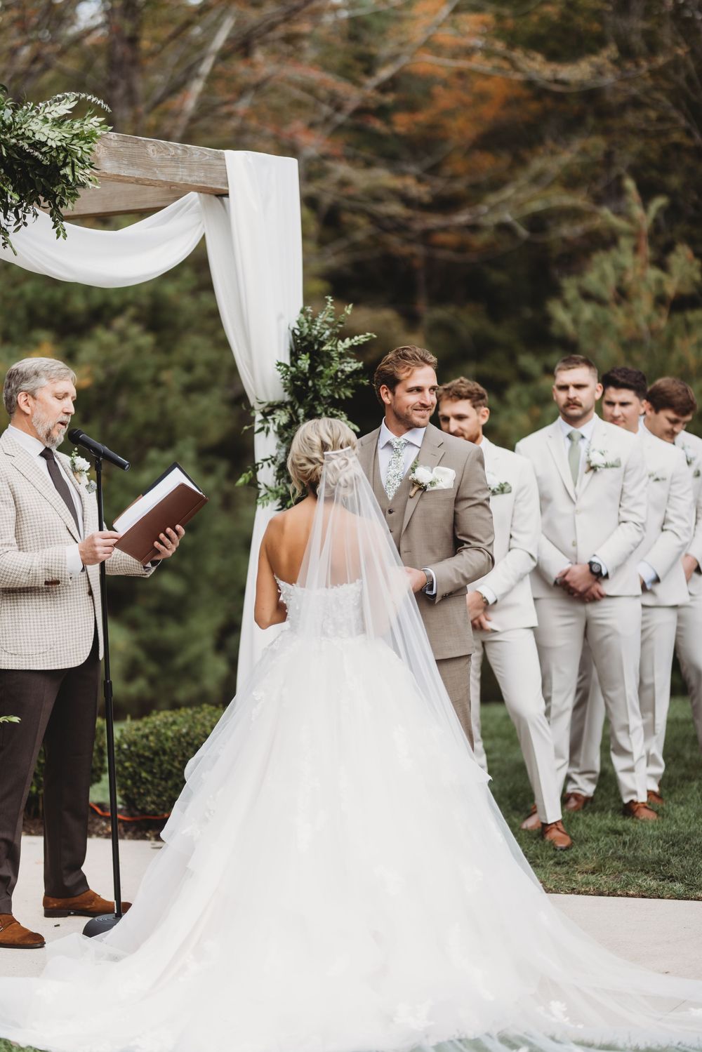 Wedding party stands outdoors during ceremony with groomsmen in light suits and bride in flowing white gown.