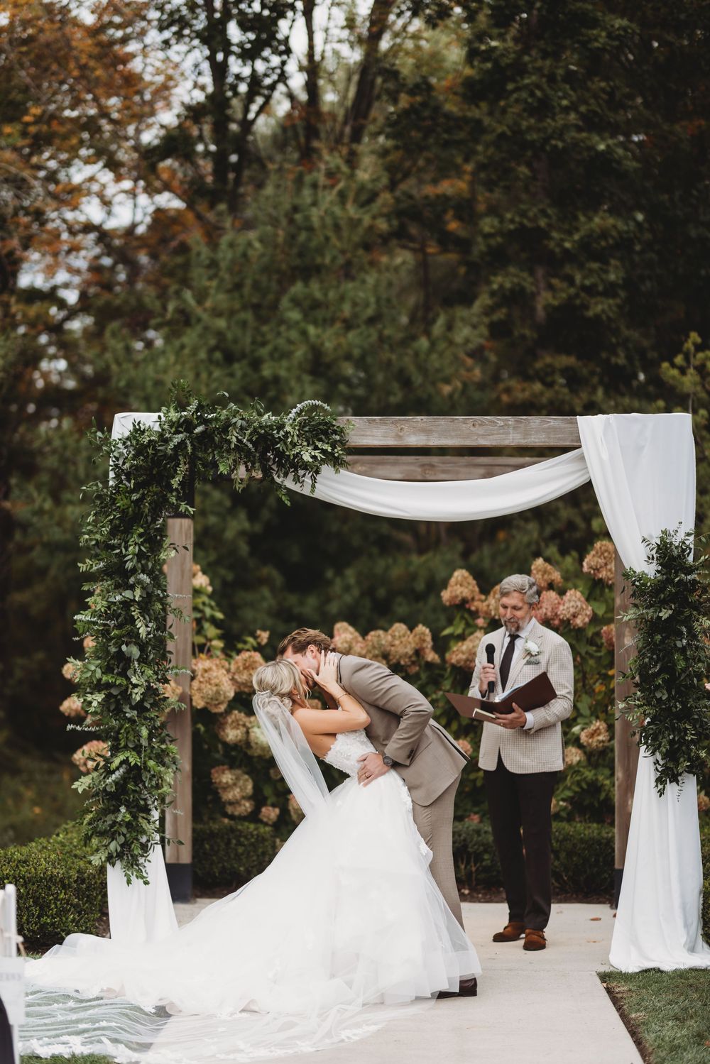 Romantic outdoor wedding ceremony setup with white fabric draping and greenery decorations in a garden setting.