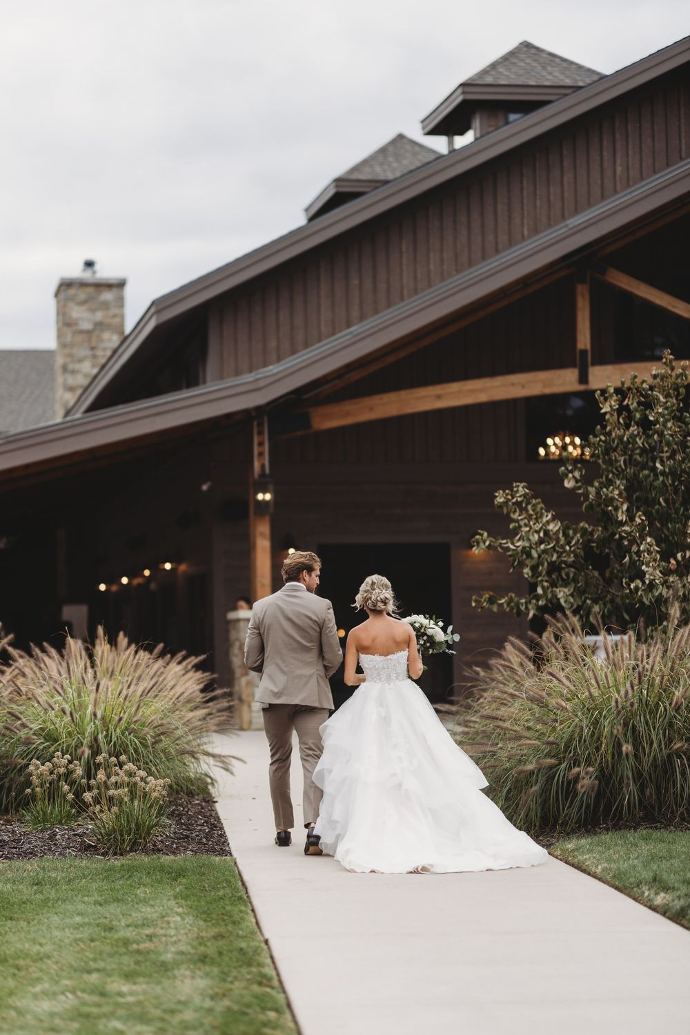 A couple in wedding attire walks along a pathway towards a rustic barn venue with ornamental grasses on a cloudy day.