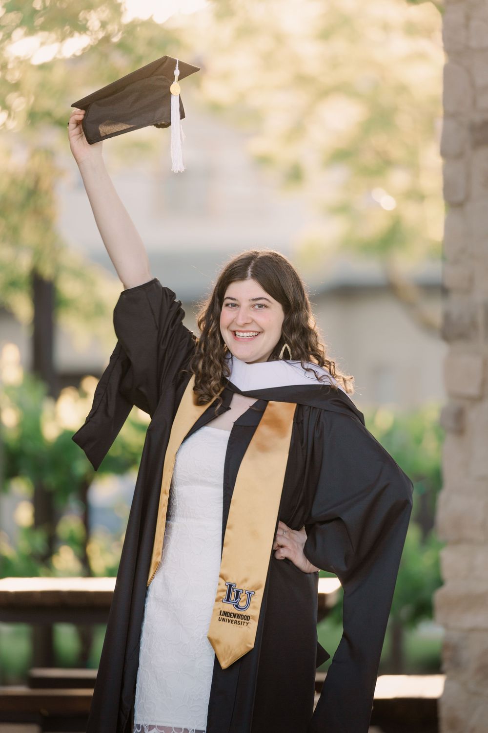A graduate in cap and gown raises their arms in celebration outdoors on a sunny day.