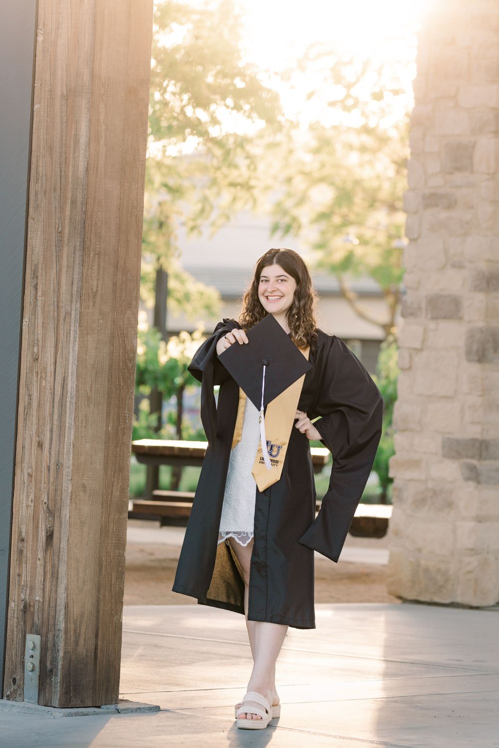Graduate in black cap and gown poses under concrete pillars at sunset during graduation photo session.