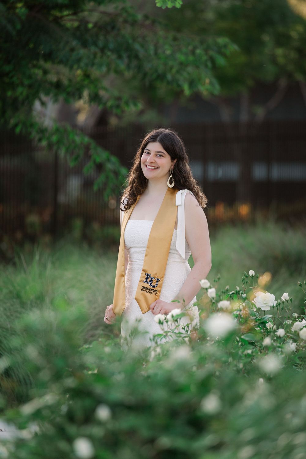Graduate in white dress and gold stole poses among blooming white flowers in a garden setting.